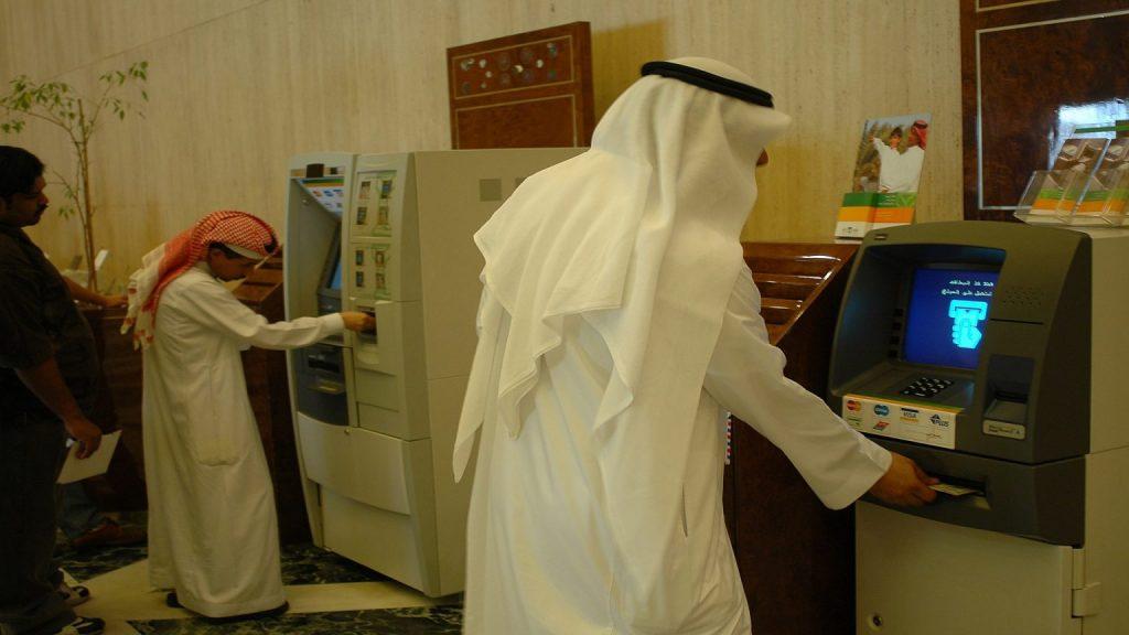 Muslim pilgrim withdrawing cash from ATM in Makkah with Kaaba in background, representing banking and money guide for Umrah pilgrims in Saudi Arabia.